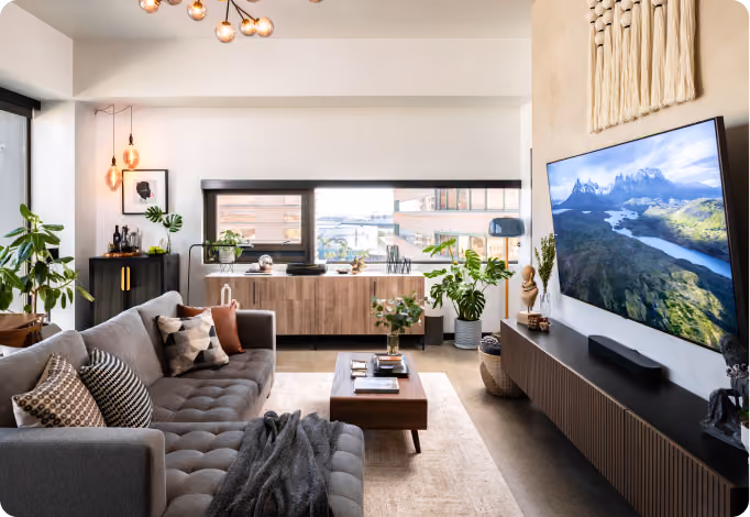 Modern living room with gray sectional sofa, wooden furniture, plants, and a large flat-screen TV on the wall.
