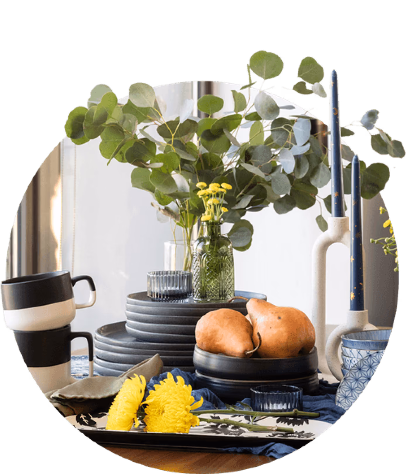 Table setting with black bowls, pears, yellow flowers, stacked black and white mugs, blue candles, and green foliage.