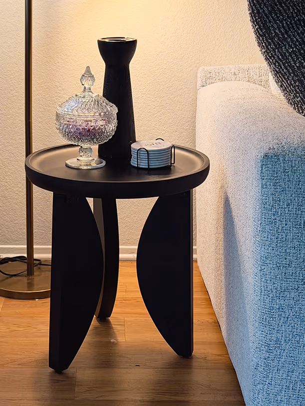 Round black three-leg side table styled with a cut-glass candy dish, matte black vase, and coasters next to a cream sofa