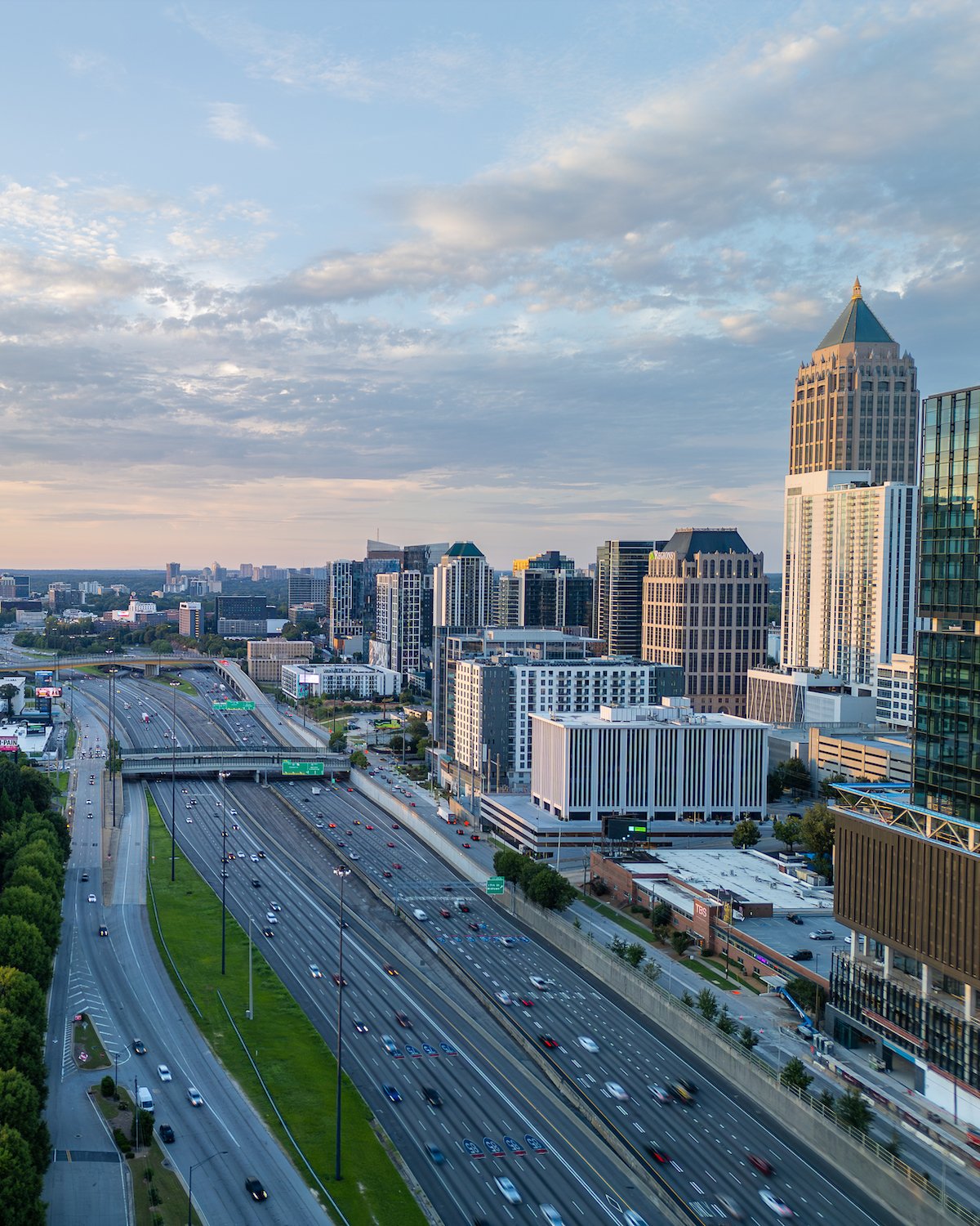 Aerial view of a multi-lane highway passing through a cityscape with tall buildings under a partly cloudy sky at sunset.