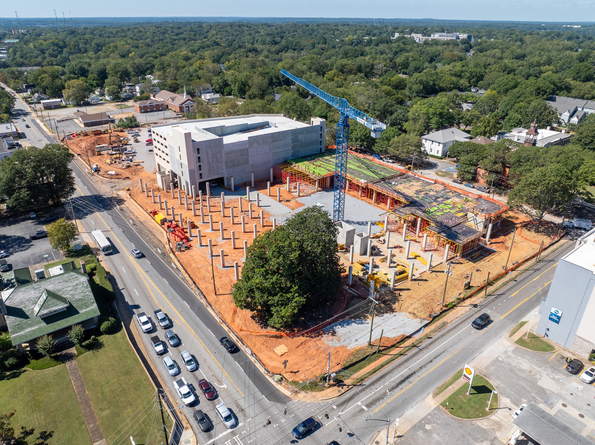 Aerial view of a large construction site with a blue crane, concrete pillars, and a partially built multi-story building surrounded by roads and trees.