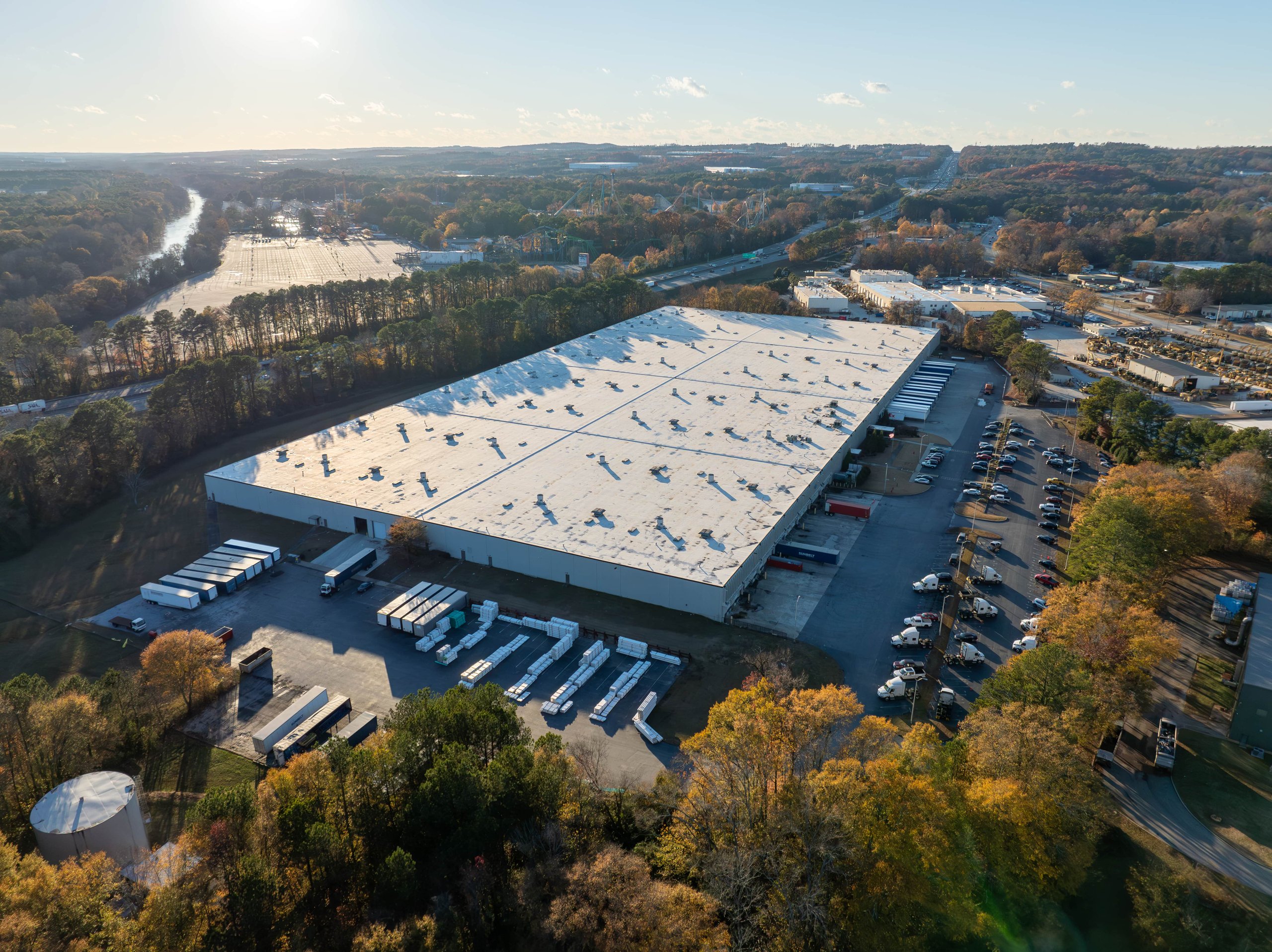 Aerial view of a large industrial warehouse with parked trucks and trailers surrounded by trees and roads in a commercial area.