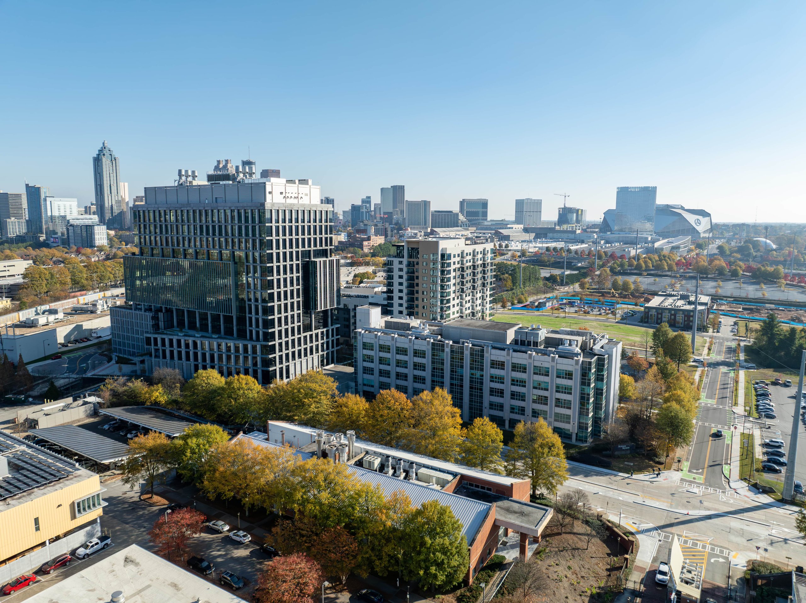 Aerial view of a cityscape featuring modern office buildings, roads, parking lots, and trees with autumn foliage under a clear blue sky.