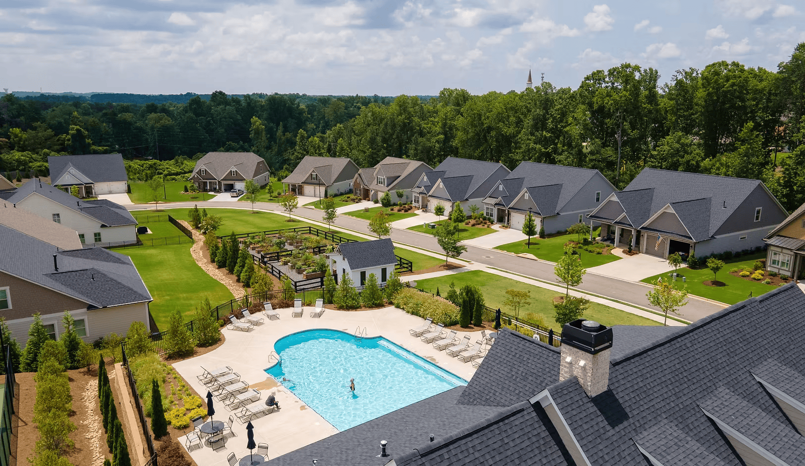 Aerial view of a residential neighborhood with houses, a fenced community swimming pool, and green lawns under a partly cloudy sky.