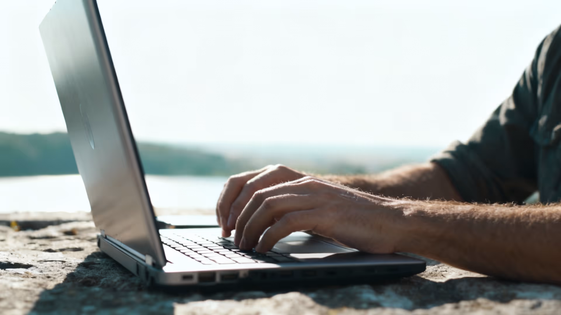 Person typing on a laptop outdoors on a stone surface with a blurred natural background.