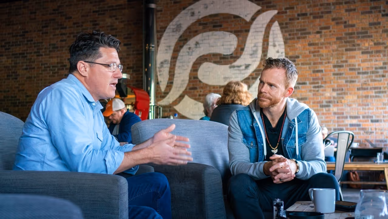 One of the founders of Enduring Legacy Mentors and a client engaged in conversation while seated in a cozy café with brick walls and a large wall mural behind them.