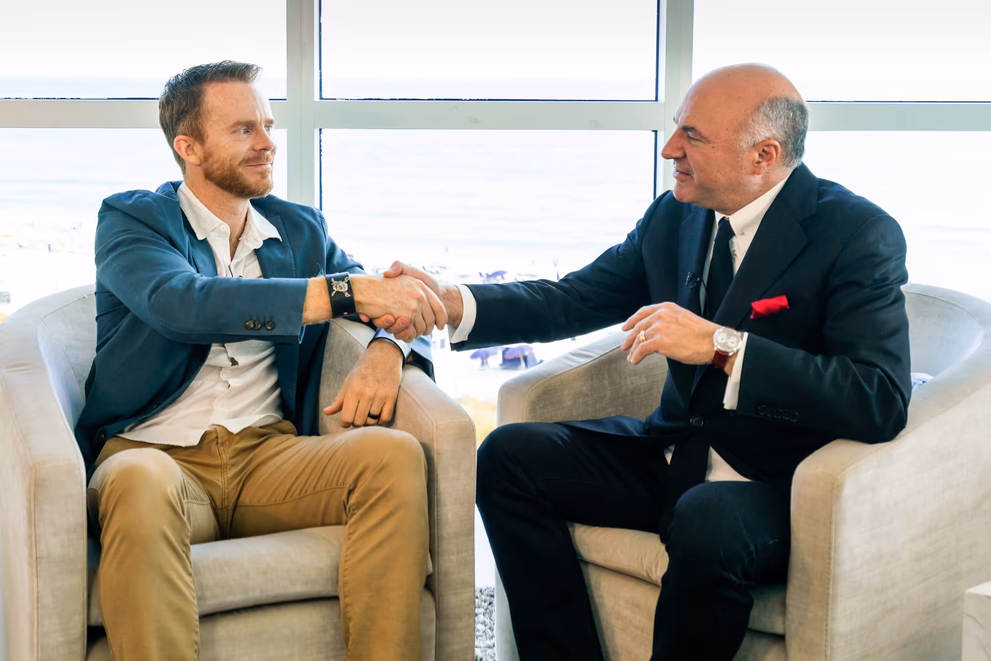 Two men sitting in armchairs shaking hands in a bright room with large windows showing an outdoor view.