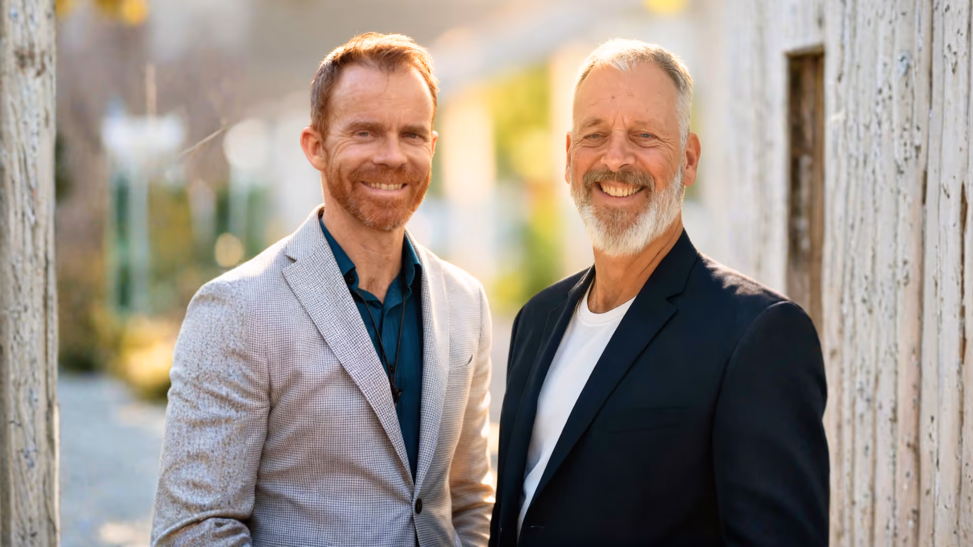 Two smiling men, one with red hair and beard in a light gray blazer and teal shirt, and the other with gray hair and beard in a black blazer and white shirt, standing outdoors near a weathered wooden wall.