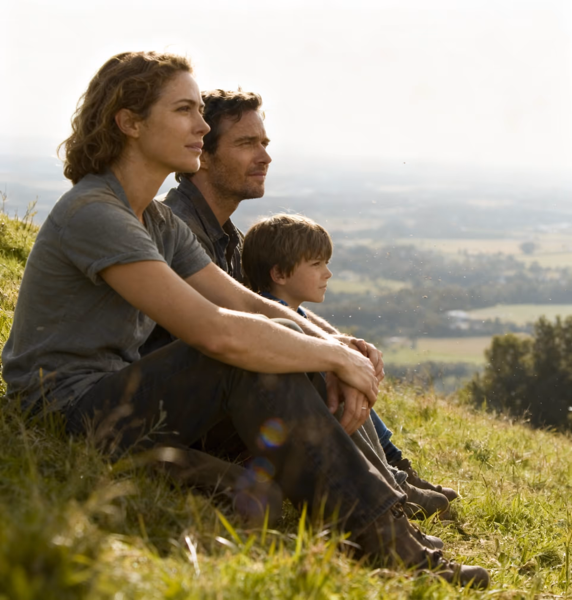 A woman, man, and boy sitting outdoors on grass, looking thoughtfully at a distant landscape.
