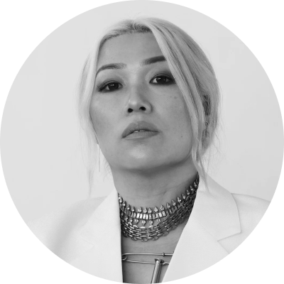 Black and white portrait of a woman with light hair wearing a white blazer and layered metallic necklace.
