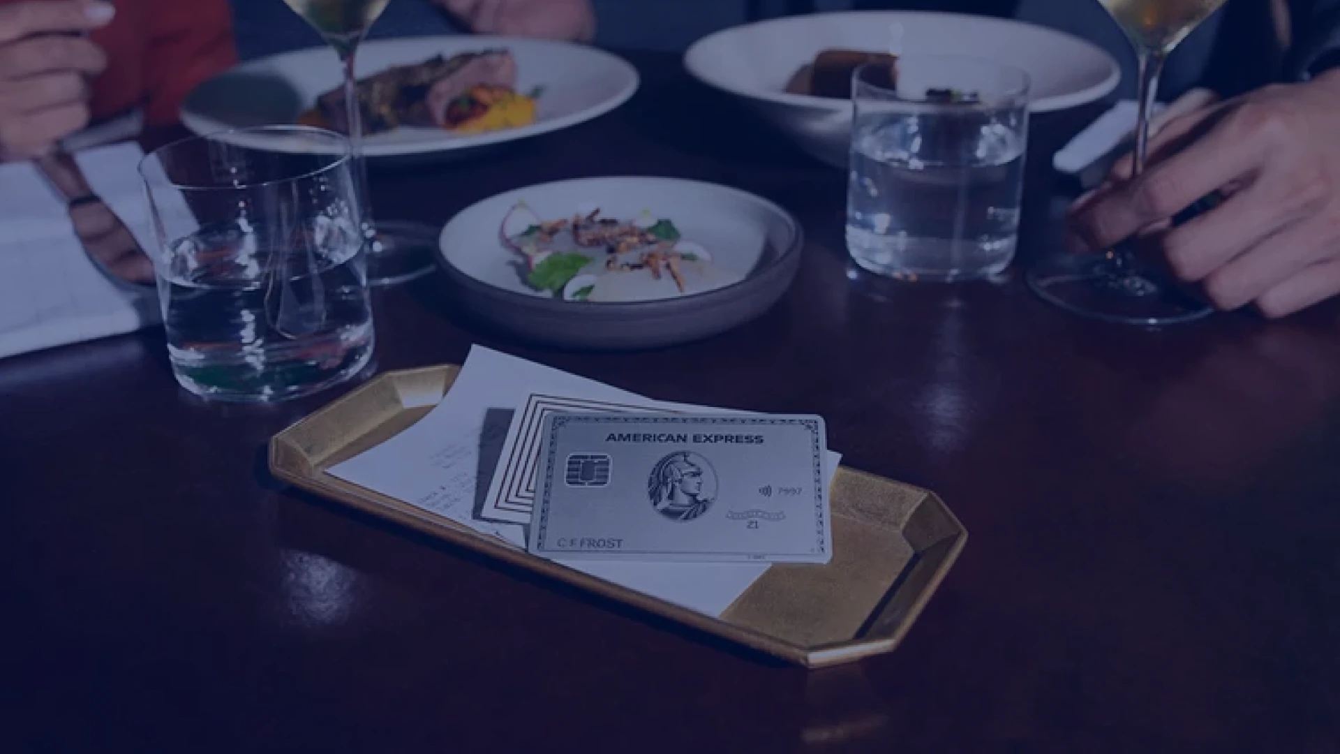 American Express card and a receipt on a tray on a restaurant table with plates, glasses, and hands in the background.