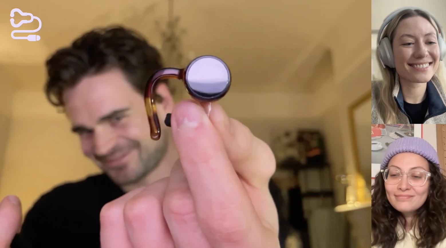 Man holding a small hearing aid device towards the camera during a video call with two smiling women.
