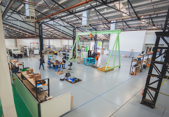Wide view of a large industrial workshop with workbenches, equipment, a green gantry crane, and two workers.