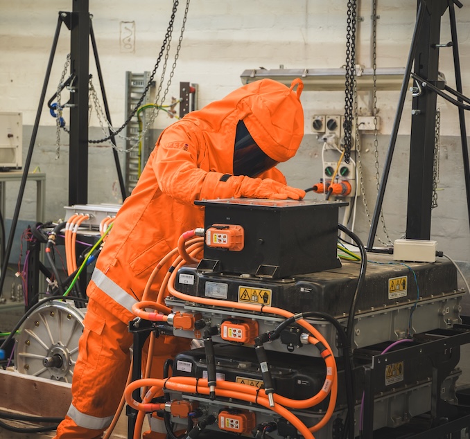 Person in an orange protective suit and hood working on industrial battery equipment with orange cables in a workshop.