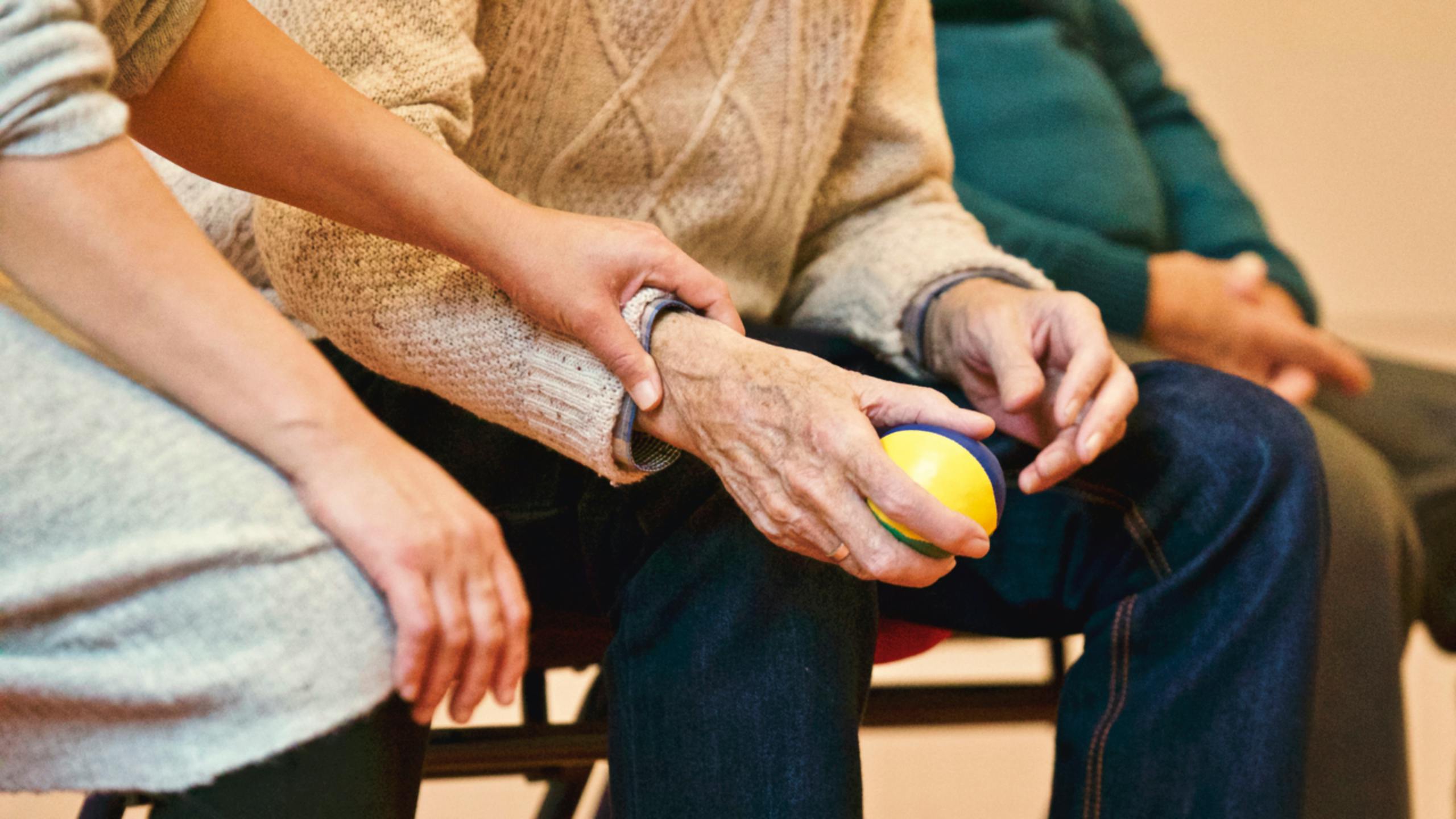 Close-up of a younger person holding the hand of an elderly person who is holding a small yellow and blue ball.