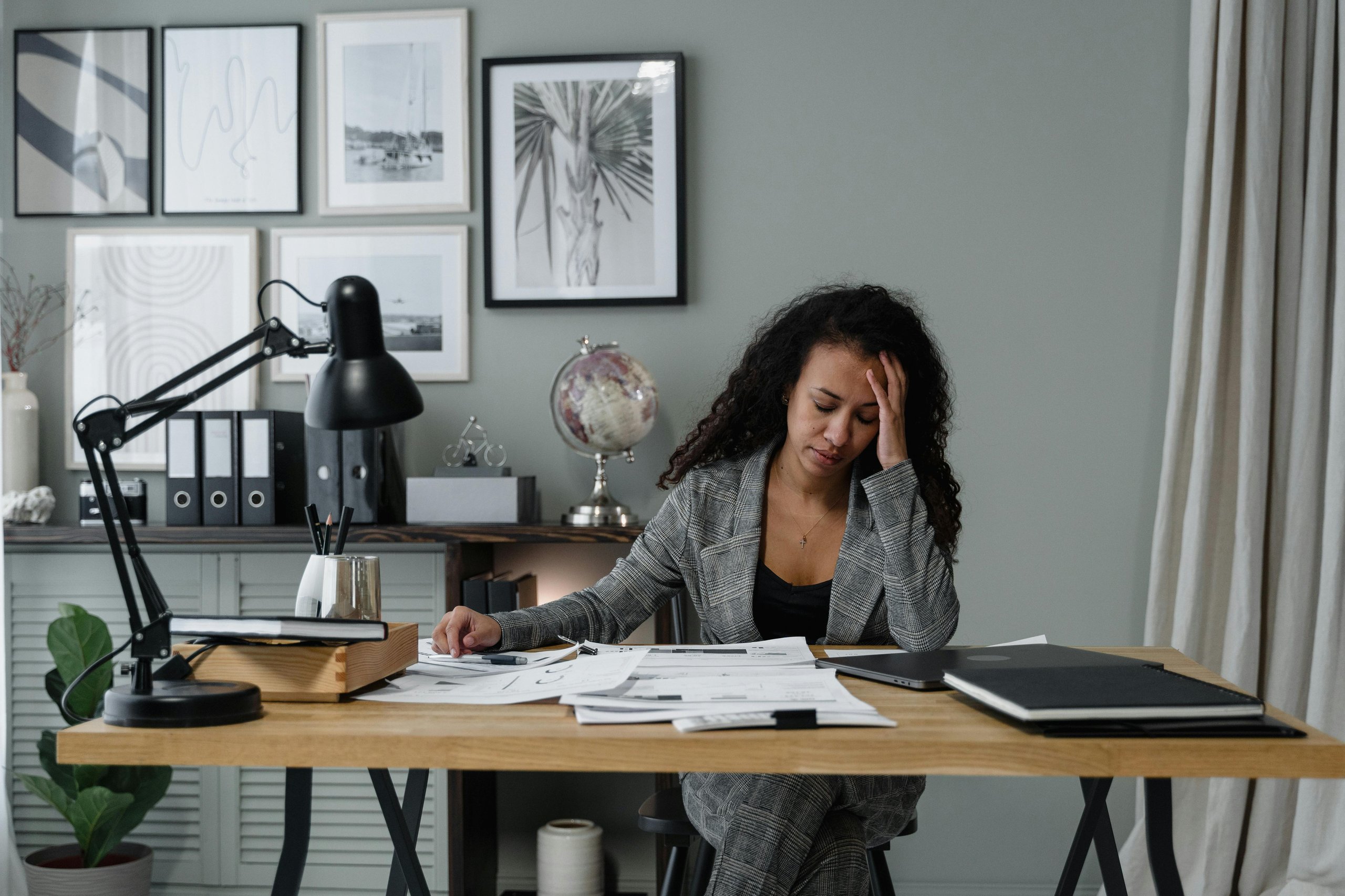 Stressed woman in a gray plaid suit sitting at a desk cluttered with papers and a laptop in a modern office.