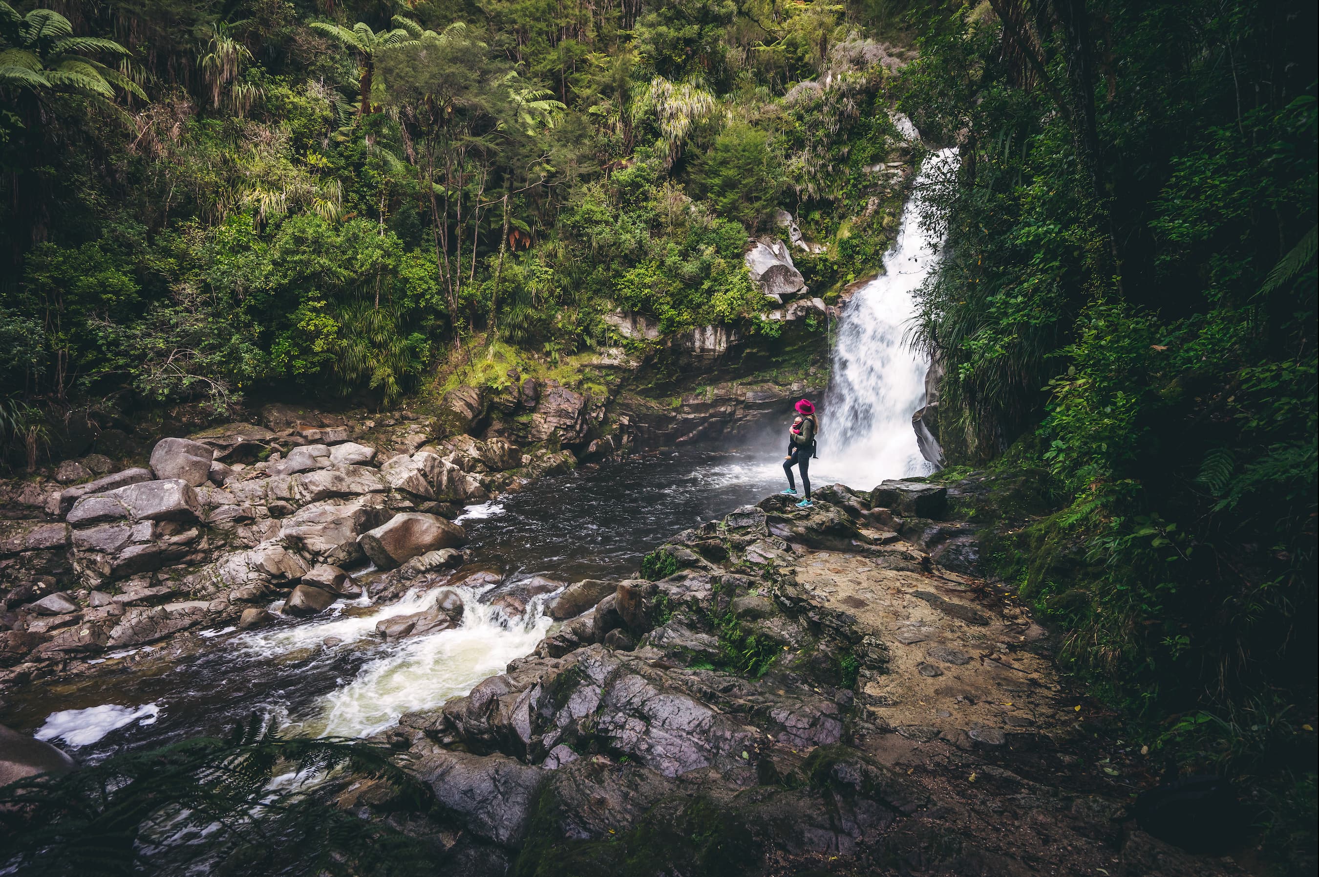 Person in a pink hat standing on rocks near a forest waterfall flowing into a river.