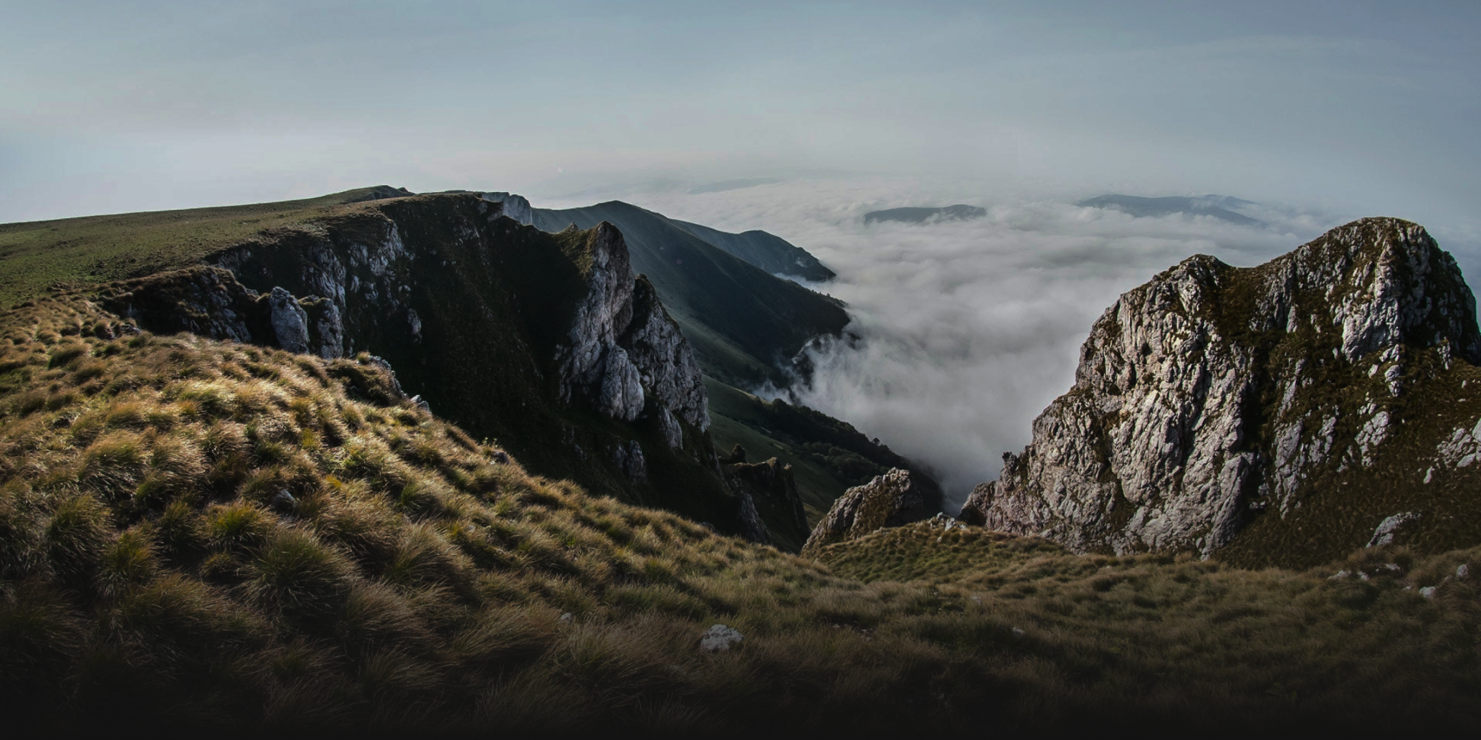 Mountain ridge with rocky cliffs and fog filling the valley below under a clear sky.