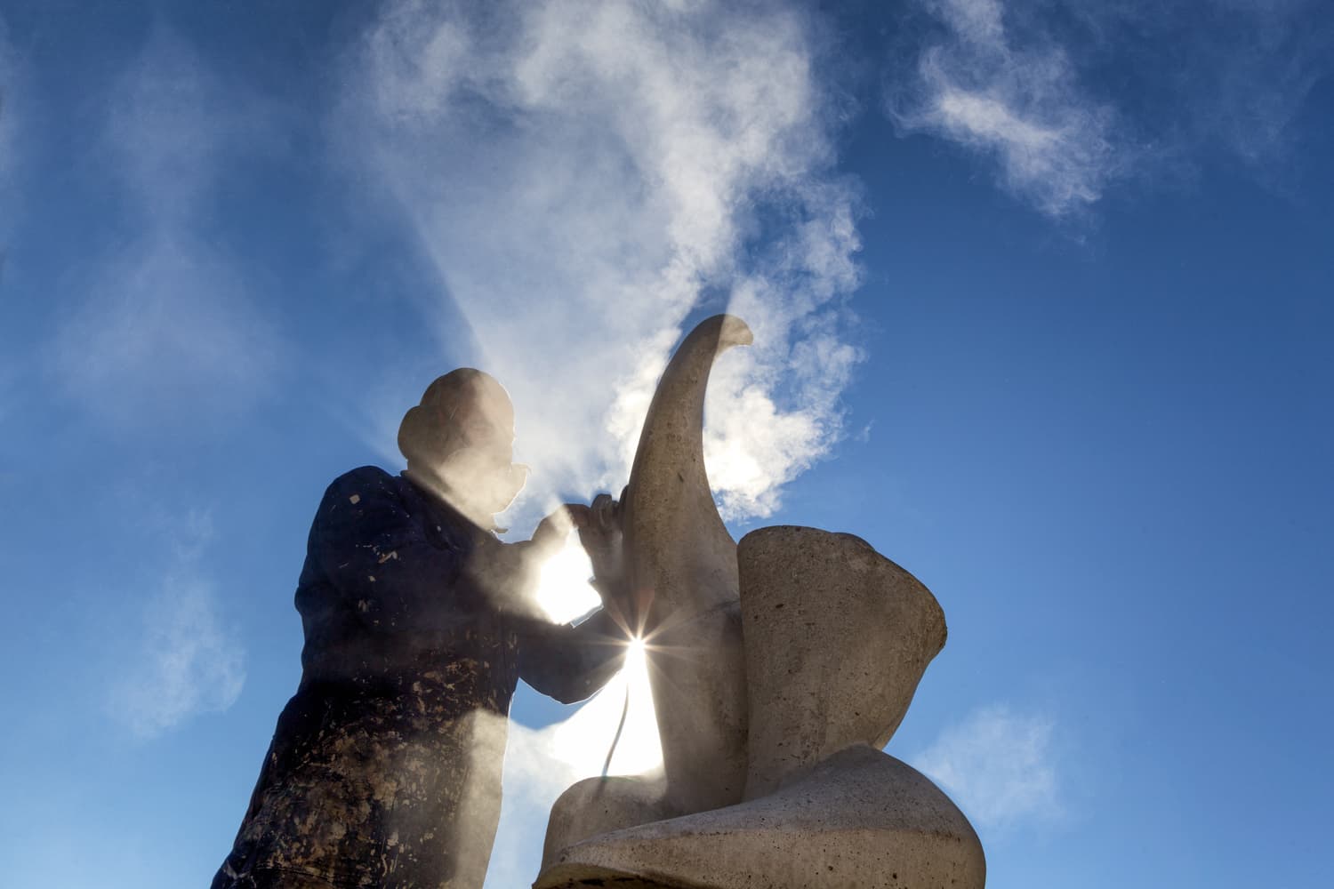Silhouetted worker cutting concrete sculpture against blue sky with sun flare and dust clouds.