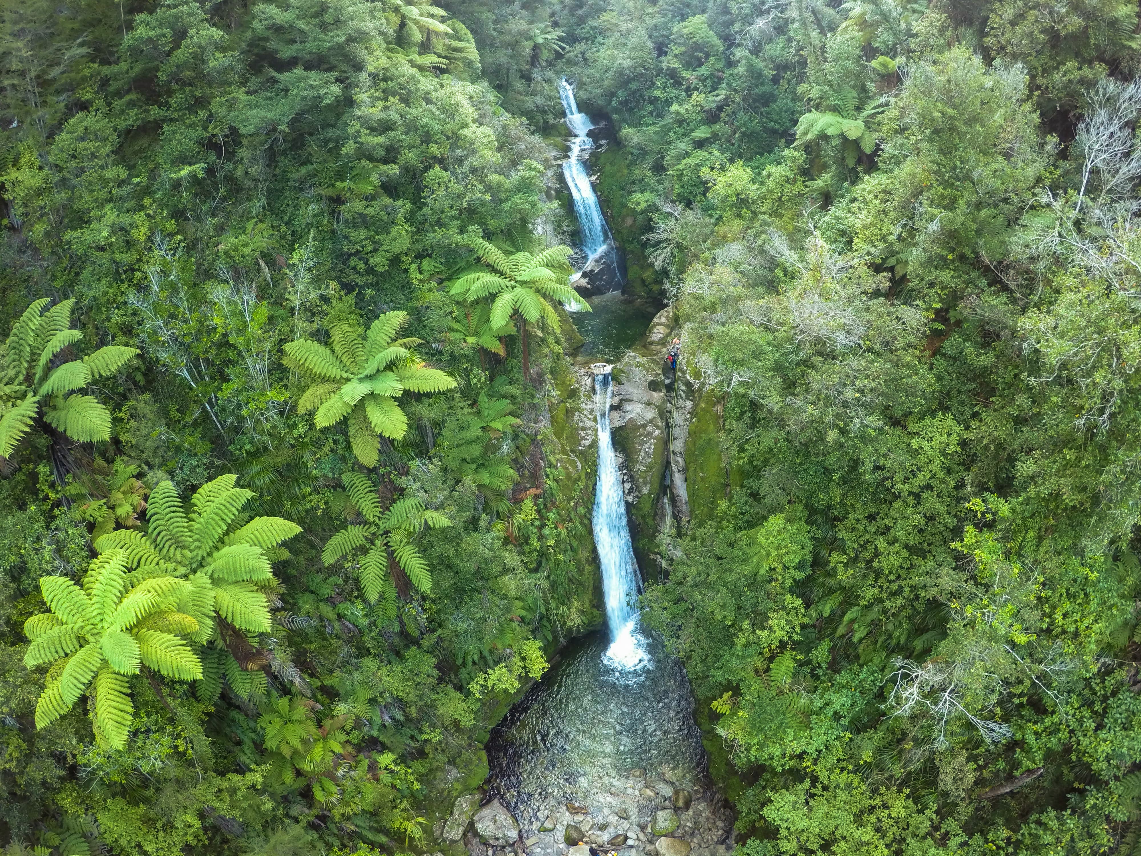 Two-tier waterfall flowing through dense green forest with a person climbing rocks beside it.