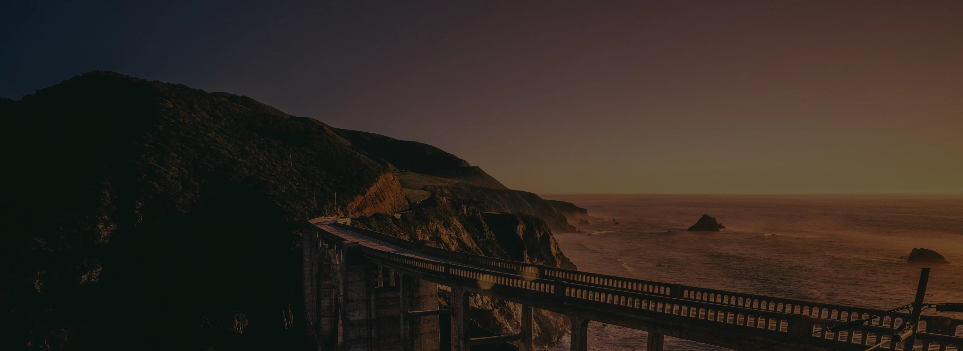 Coastal bridge extending over cliffs with ocean waves at sunset.