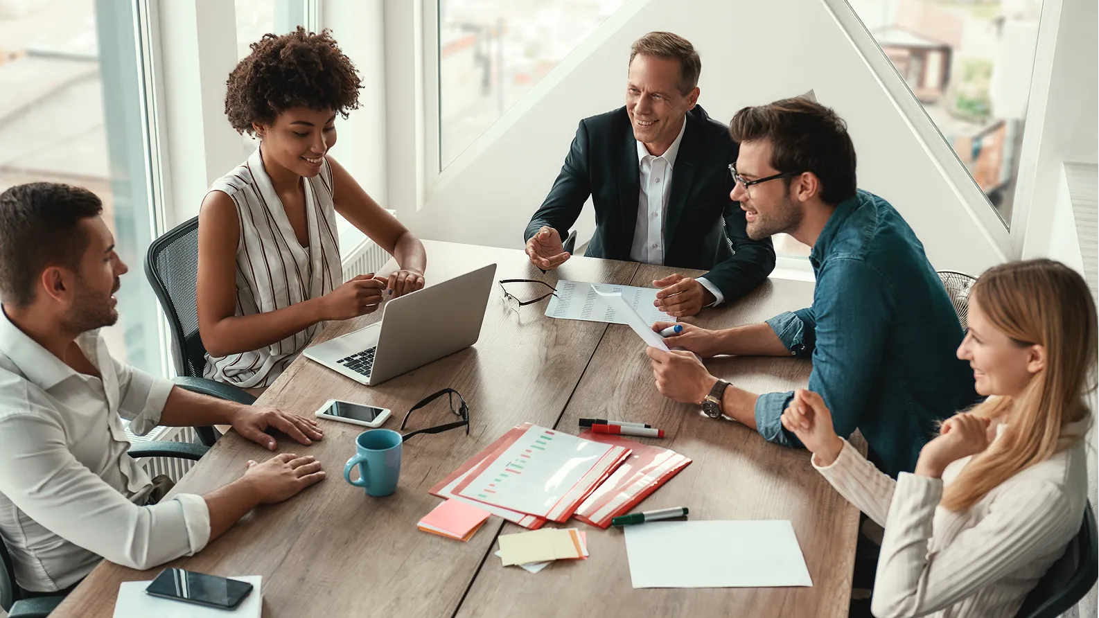 Five diverse professionals smiling and discussing work around a table with laptops, papers, and coffee mugs in a bright office.
