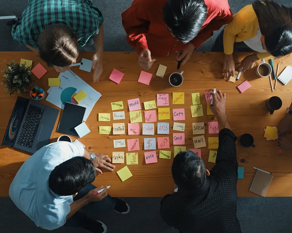 Overhead view of six people collaborating around a table covered with colorful sticky notes, laptops, notebooks, and coffee cups.