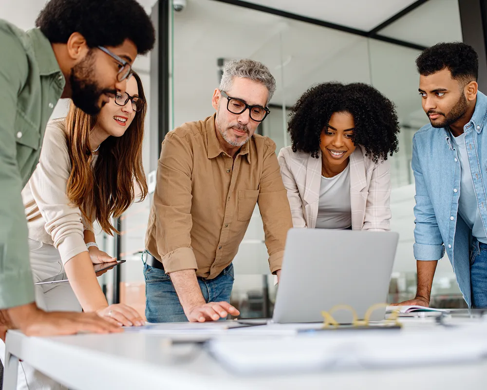 Group of five diverse professionals gathered around a table looking at a laptop in a modern office.