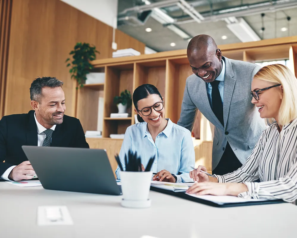 Four diverse professionals smiling and collaborating around a desk with laptop and documents in a modern office.