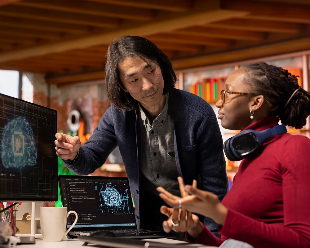 Two colleagues discussing artificial intelligence concepts displayed on a desktop monitor and laptop screen in a modern office.