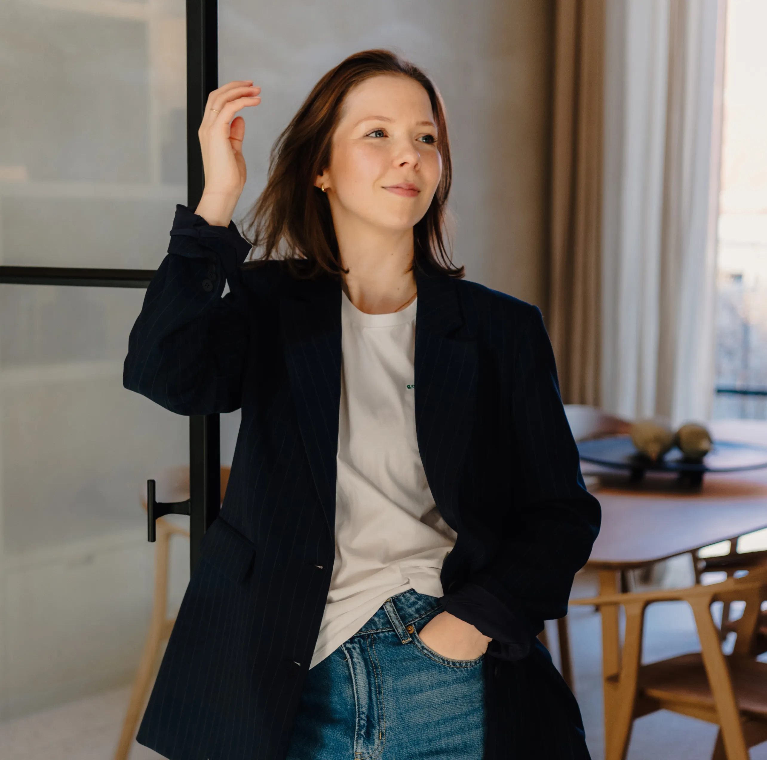 Jolien Herthogs met een donkerblazer, een wit T-shirt en een spijkerbroek staat ontspannen in een lichte kamer met houten meubels.