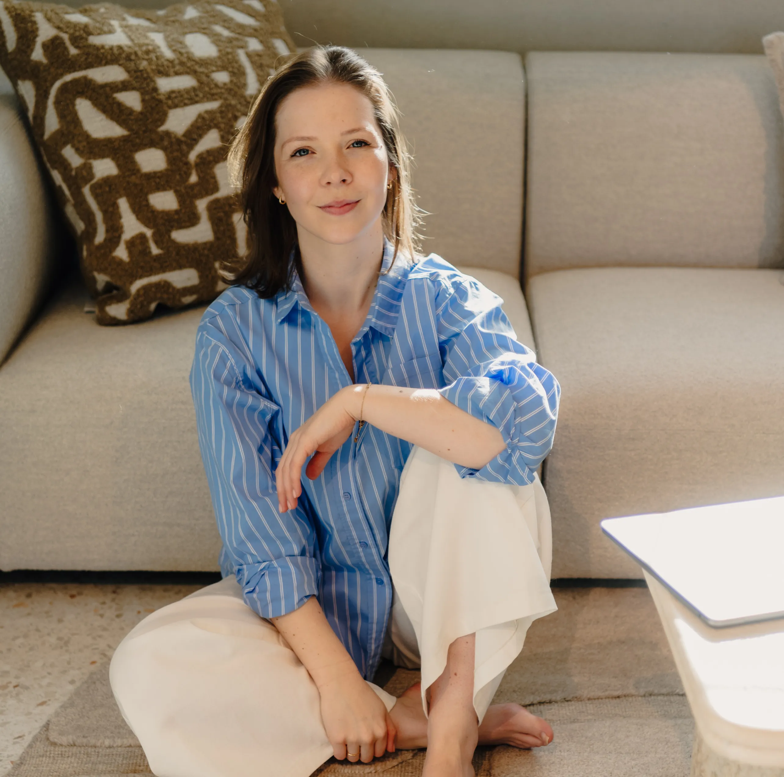 Vrouw met lichtblauw gestreept overhemd en witte broek zit ontspannen op de grond voor een beige bank met een patroon kussen.