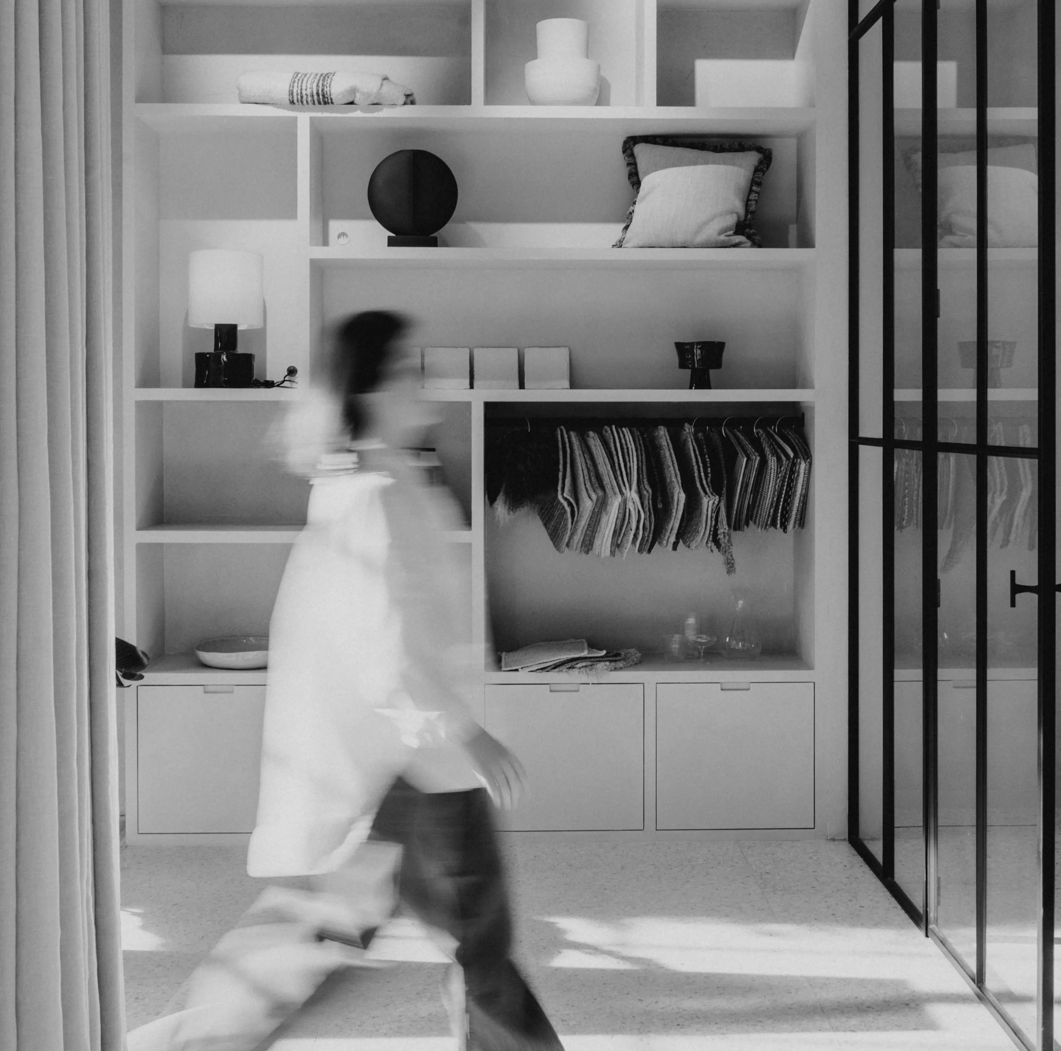 Wazig beeld van een persoon die langs een kast met opgevouwen stoffen en decoratieve items loopt in een moderne, lichte kamer.