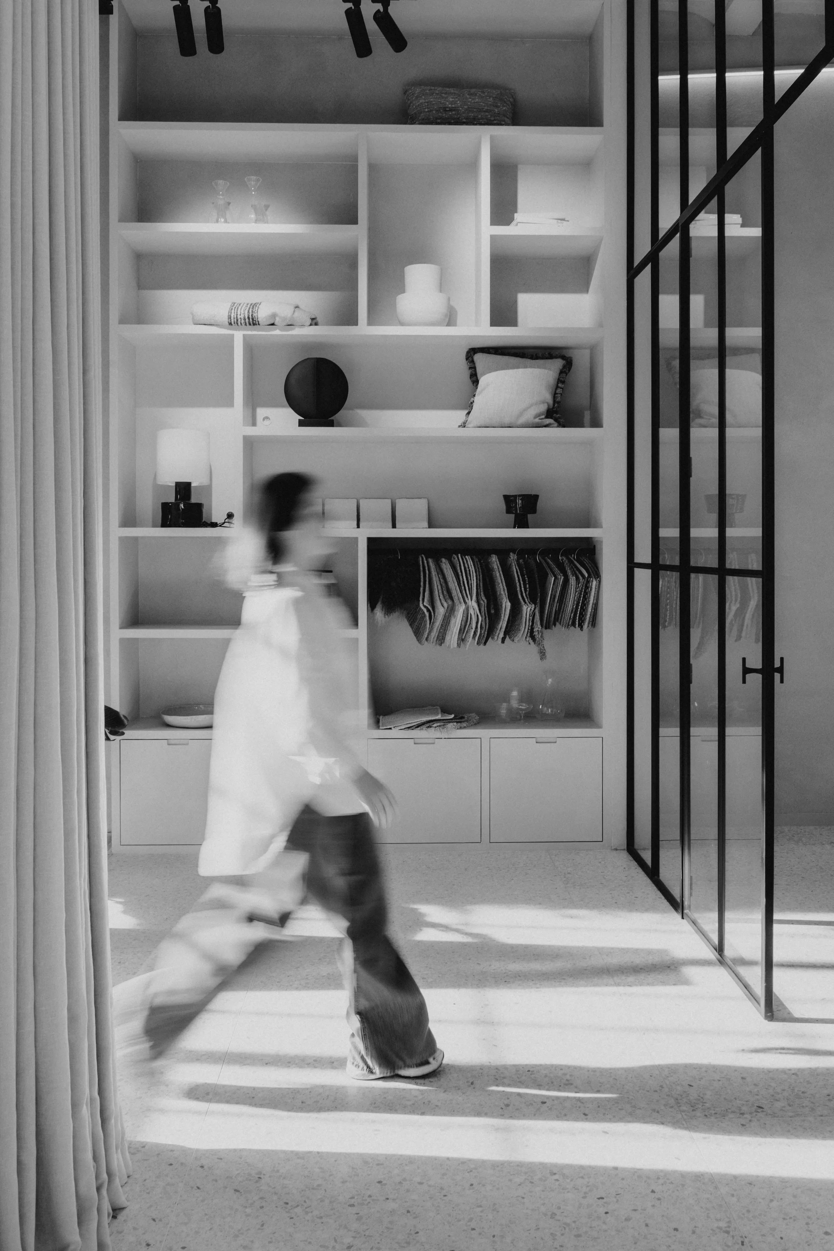 Wazig persoon loopt door een moderne kamer met wandplanken vol stoffen, kussens, en decoratie, naast een glazen deur.