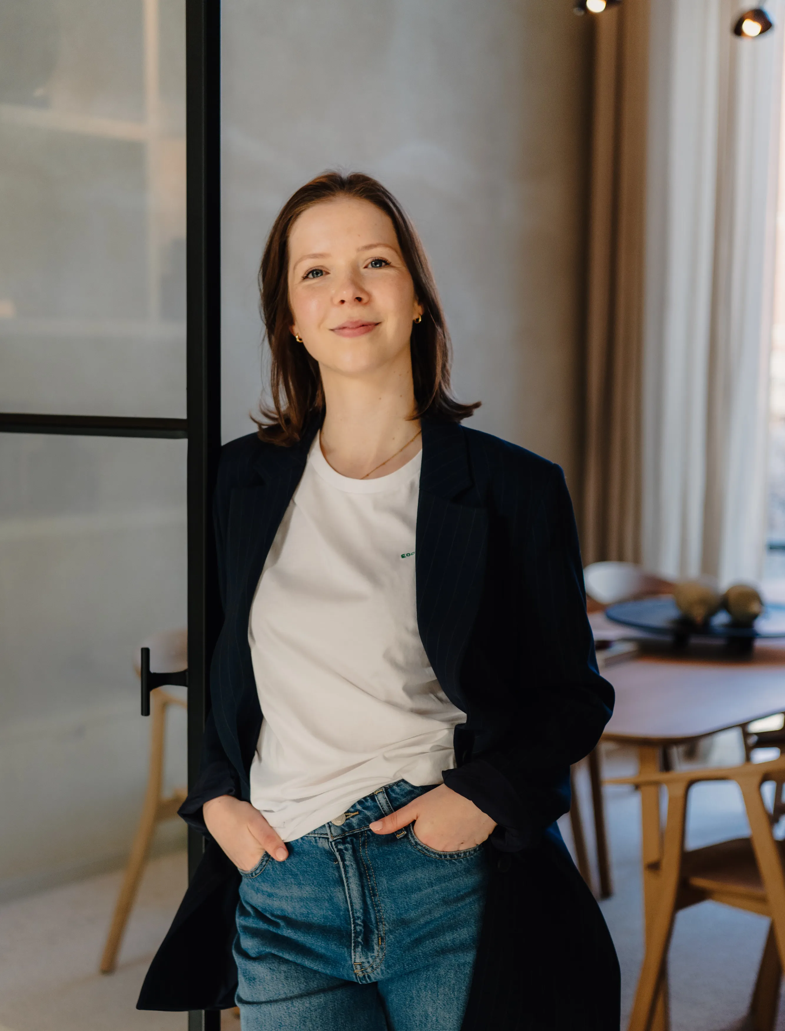 Jolien Herthogs met een wit T-shirt en donker colbert staat ontspannen met handen in haar spijkerbroekzakken in een lichte kamer.