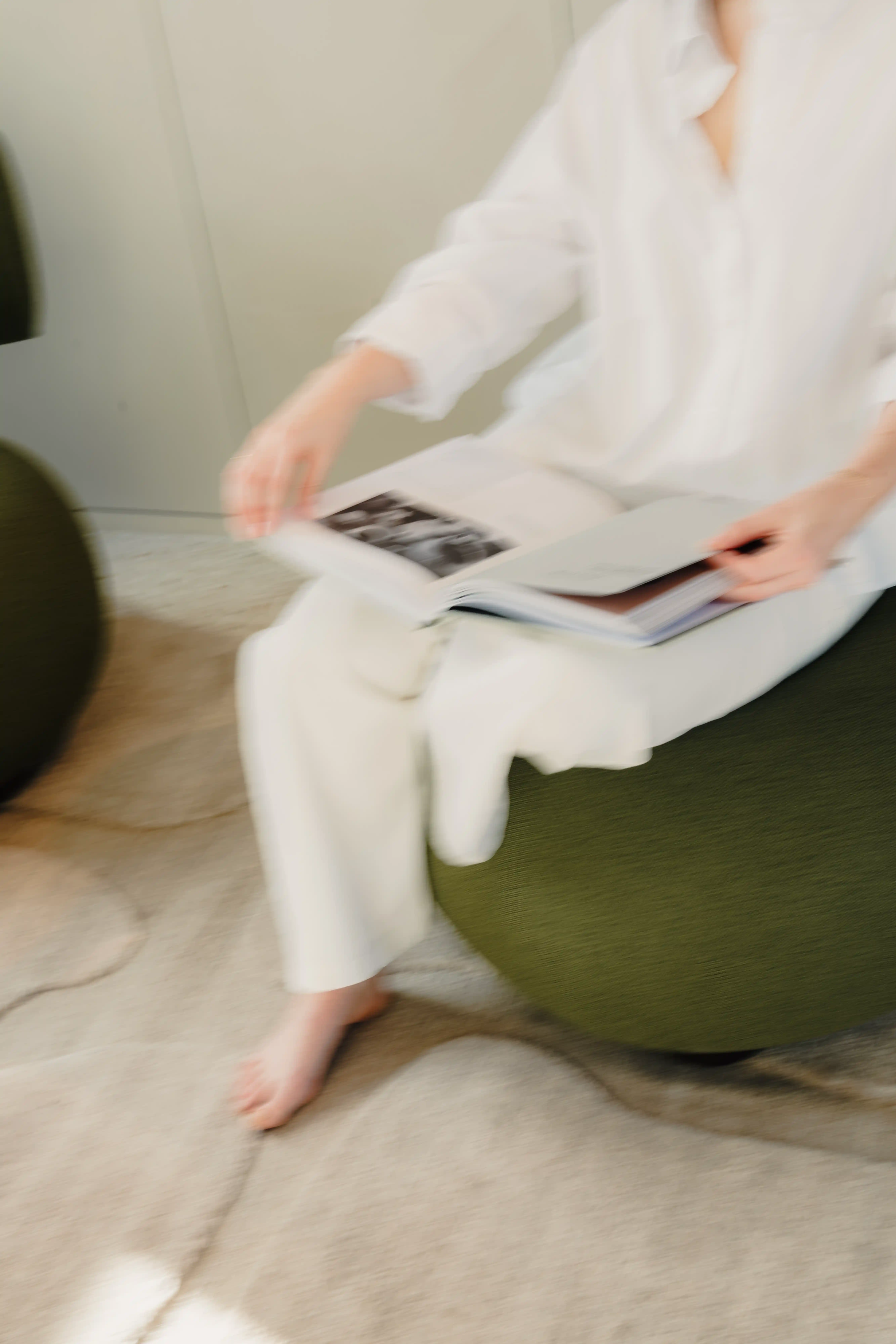Persoon in wit kleding zittend op een groene stoel en een boek bladerend met blote voeten op een lichte vloer.