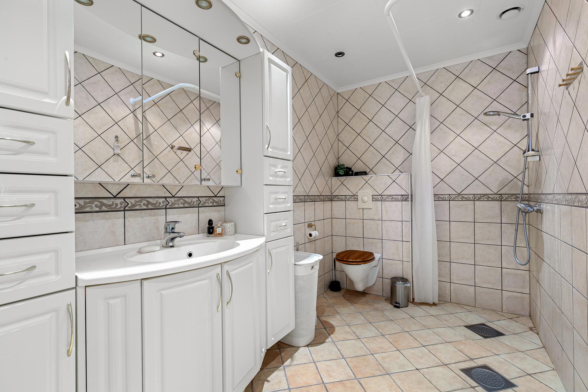Heilflisa bad med varmekablar i golv. Badet er utstyrt med servant med innreiing, vegghengt toalett og dusj.
