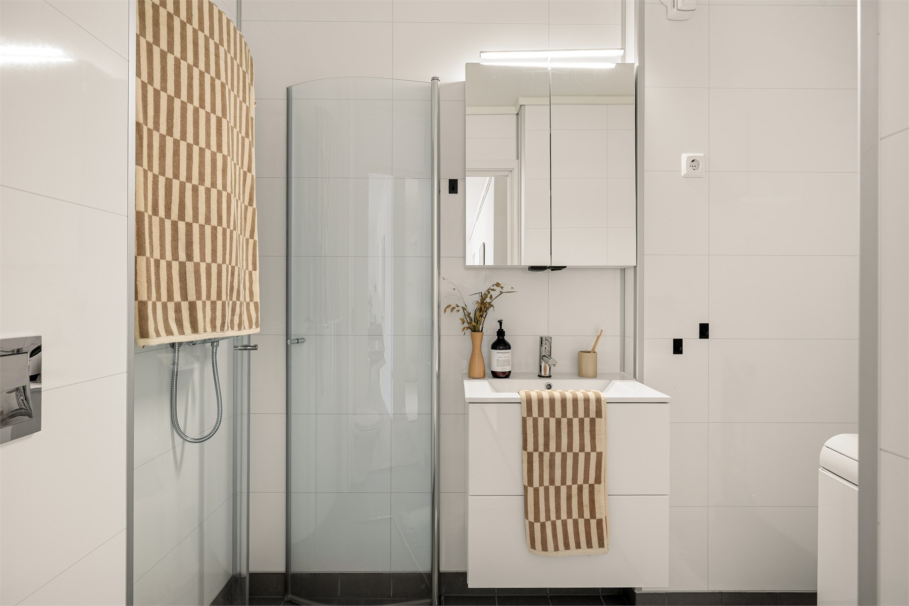 Badet kombinerer et stilrent uttrykk med praktiske løsninger som vegghengt toalett, servant med skuffer, dusjdører i klart glass og opplegg for vaskemaskin.