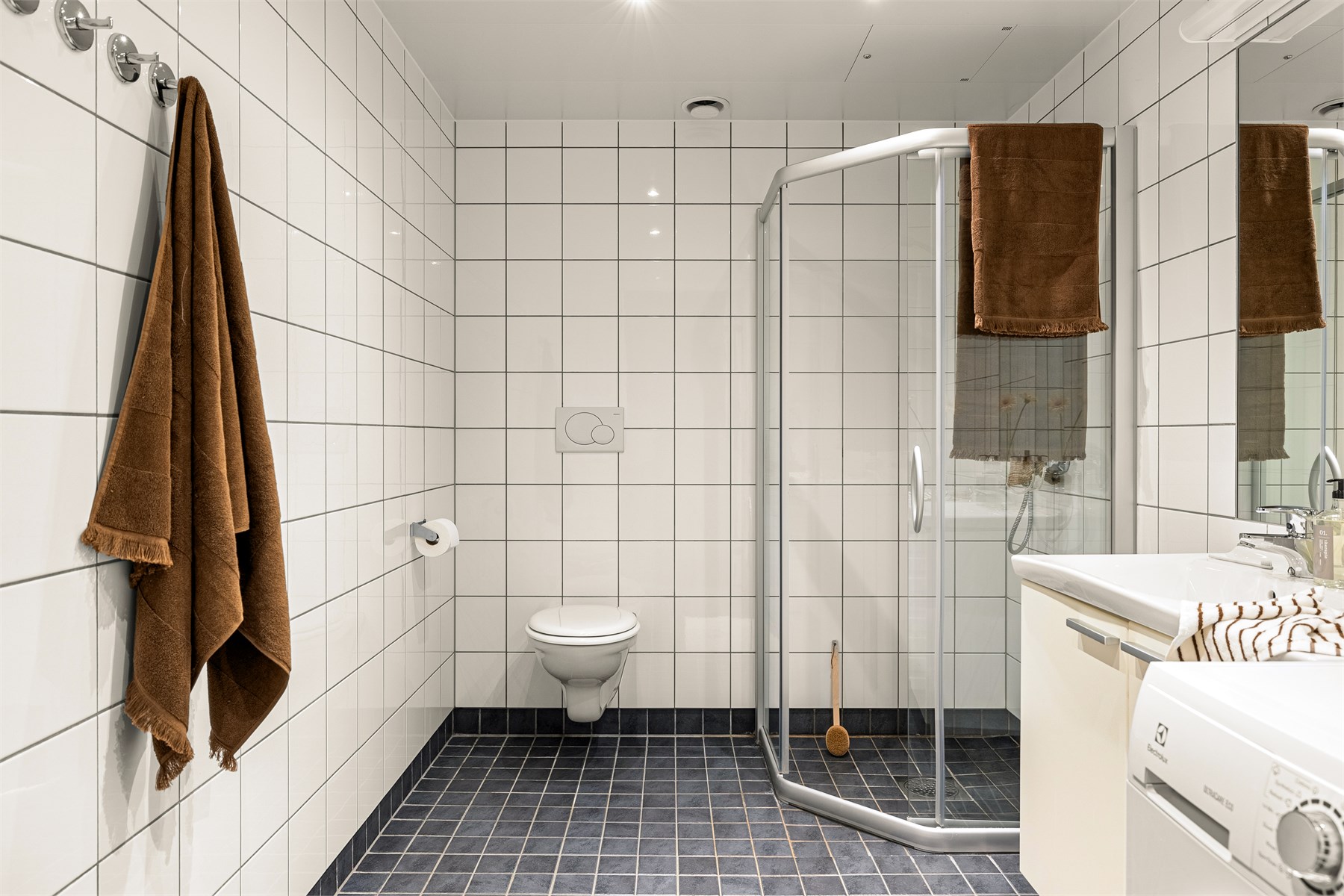 Badet er innredet med vegghengt toalett, helstøpt servant og praktisk oppbevaring i skap under servanten. Dusjdører i klart glass bidrar til å bevare den åpne romfølelsen.