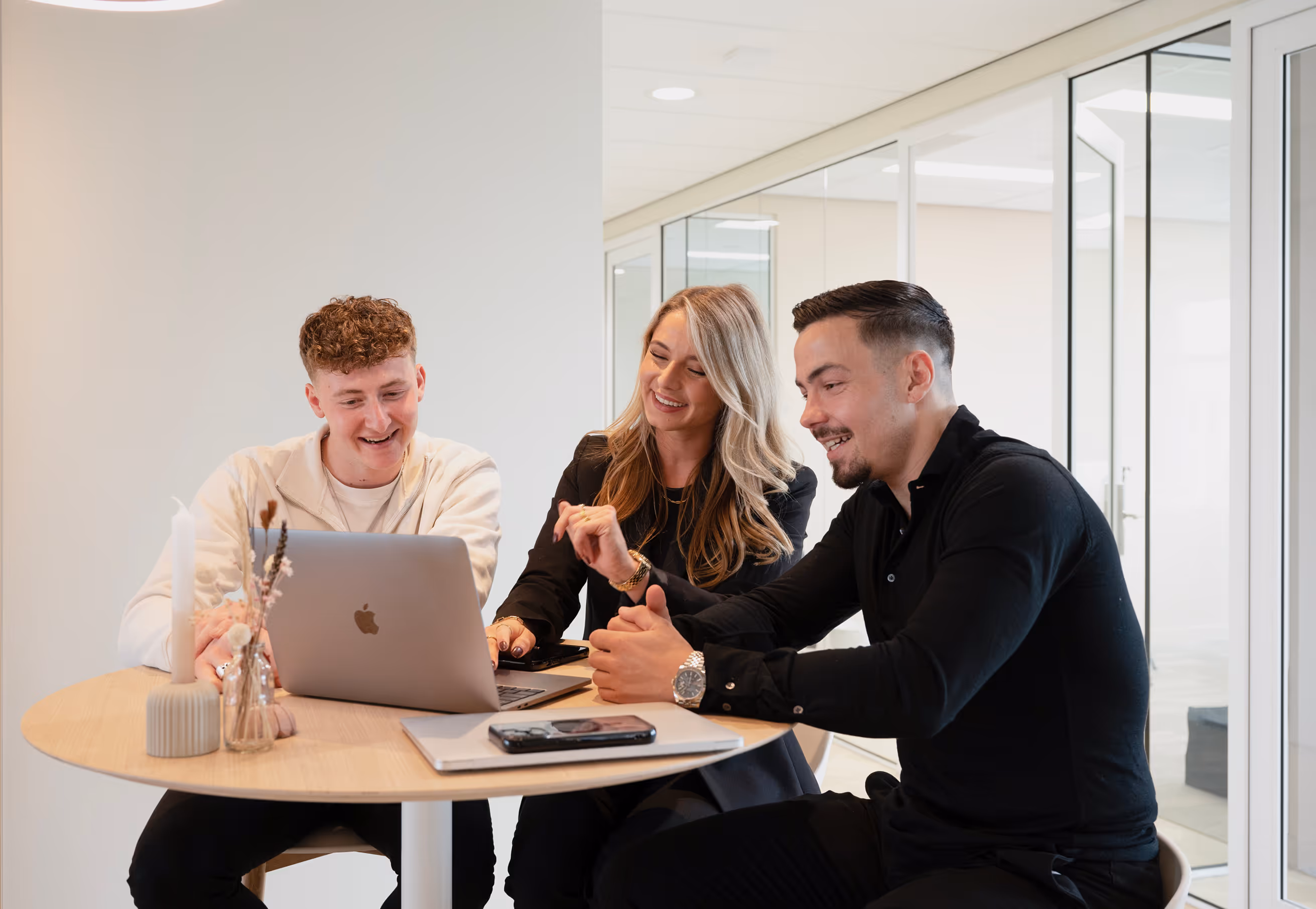 Drie jonge volwassenen lachen en werken samen aan een laptop aan een ronde tafel in een moderne kantooromgeving.