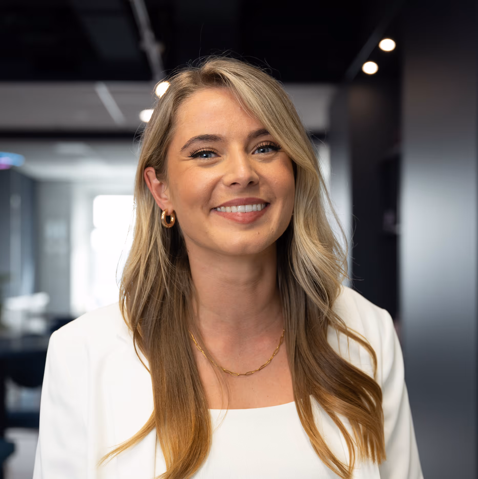Siska van topclicks. Lachende vrouw met lang blond haar, witte blazer en gouden ketting in een moderne kantooromgeving.