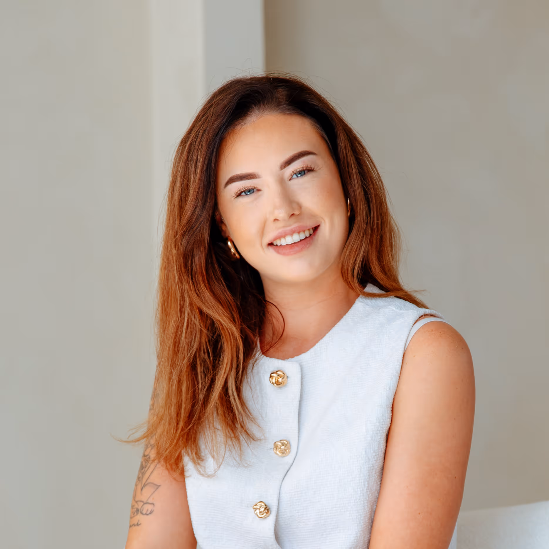 Glunderende vrouw met lang kastanjebruin haar, witte mouwloze top met gouden knopen en tatoeage op haar arm.