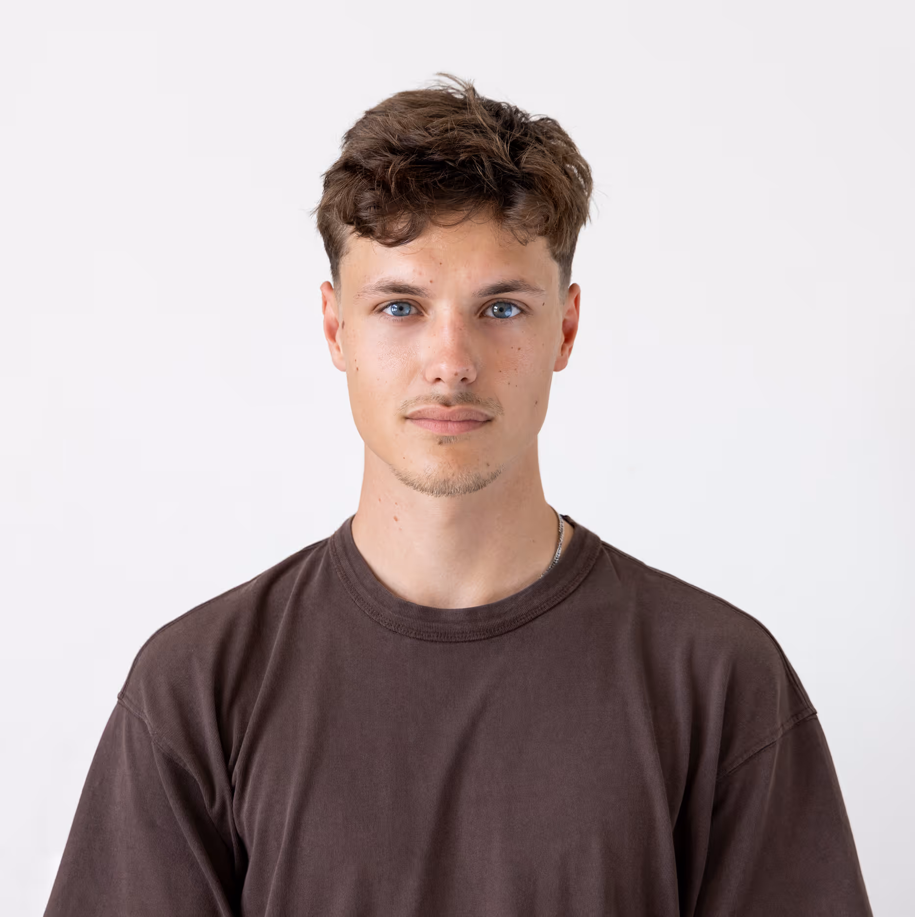 Jonge man met blauwe ogen en kort golvend bruin haar draagt een donkerbruin T-shirt, staande voor een witte achtergrond.
