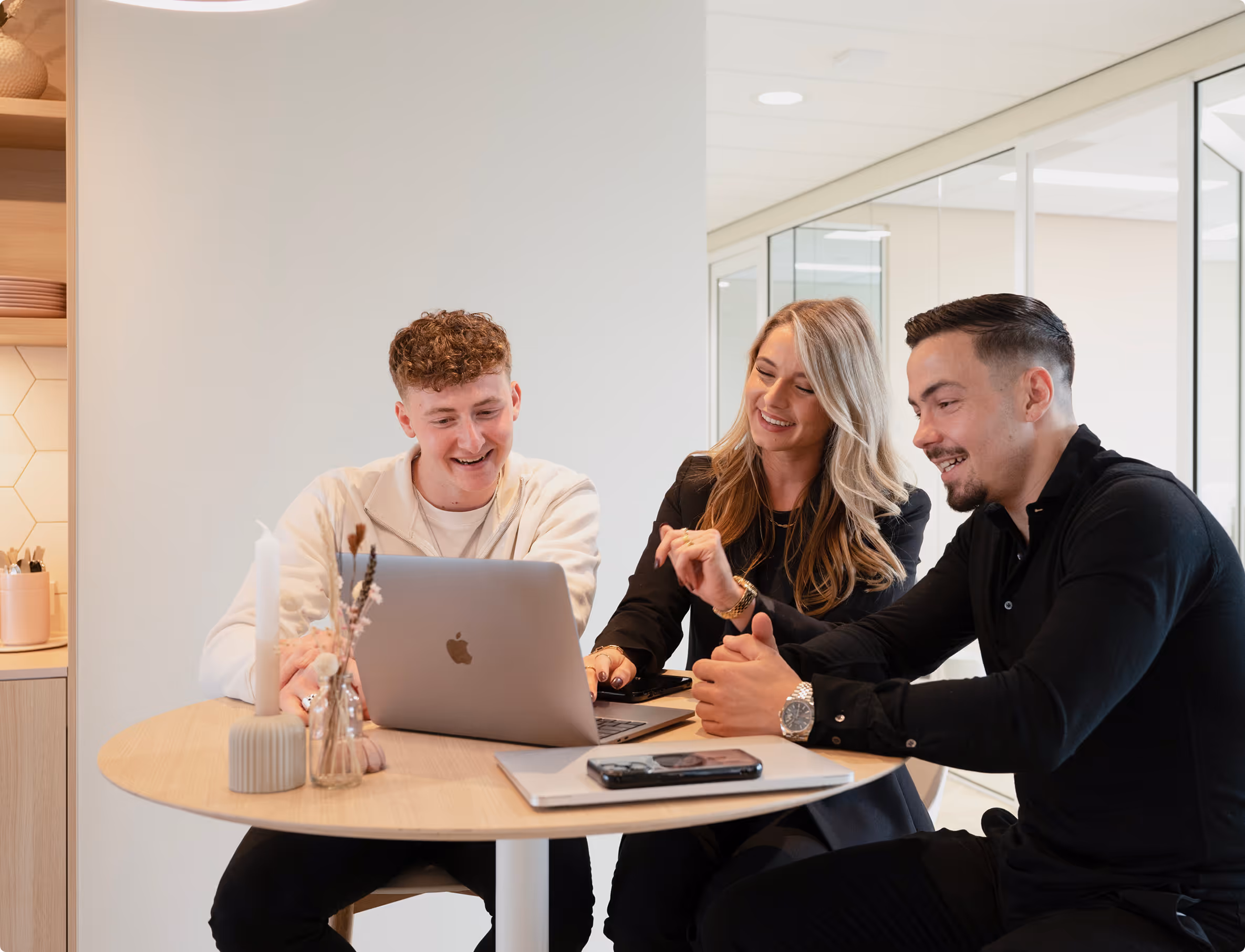 Drie jonge volwassenen die glimlachend rond een tafel zitten en samenwerken op een laptop in een moderne kantoorruimte.
