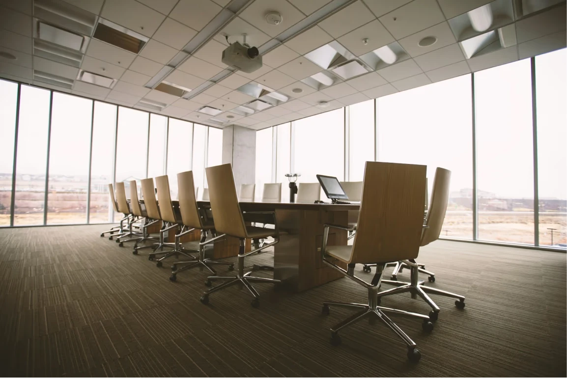 Salle de conférence moderne avec grande table entourée de plusieurs chaises à roulettes et fenêtres panoramiques laissant entrer la lumière naturelle.