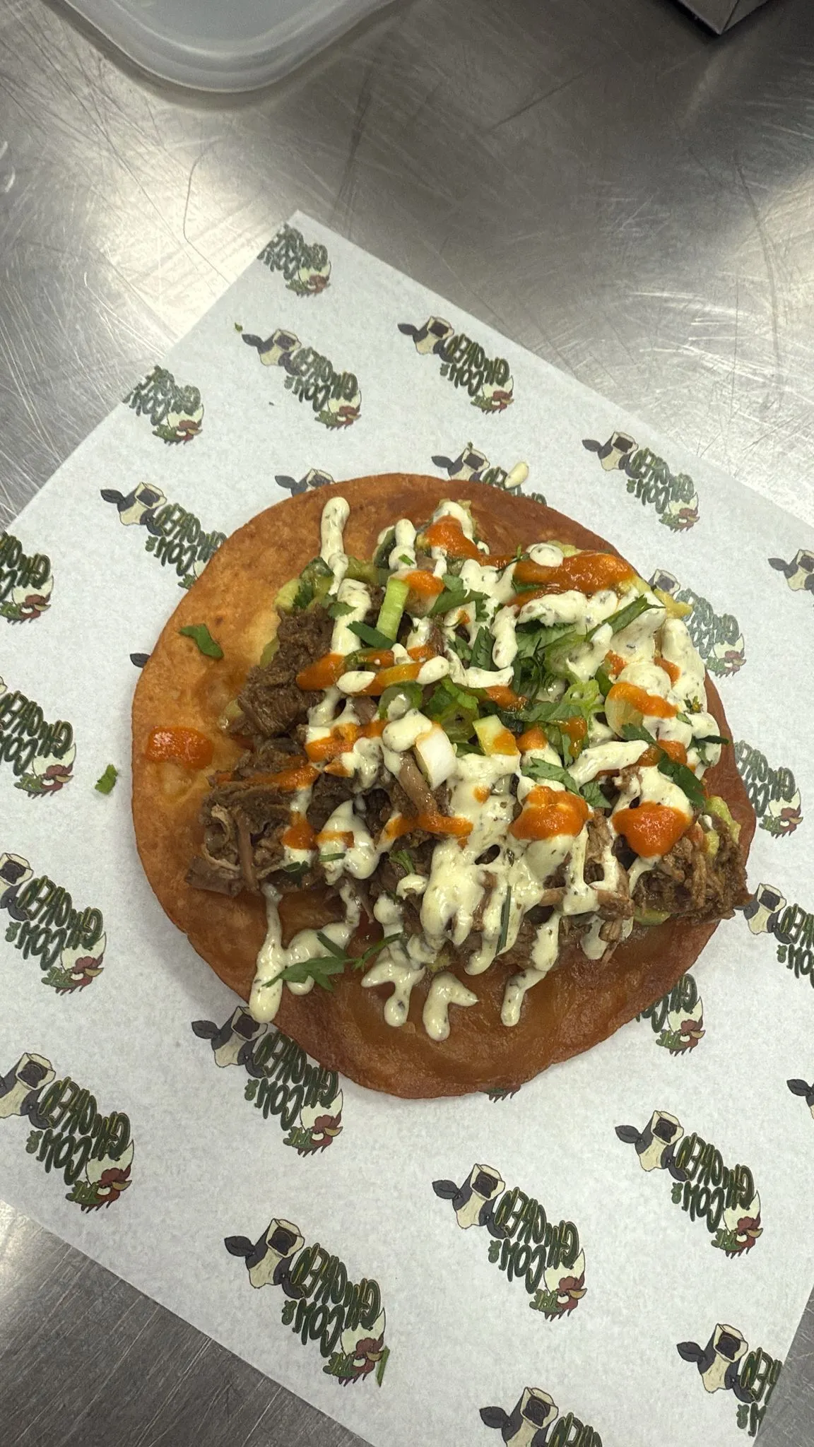 Fried taco topped with shredded meat, chopped green onions, cilantro, and drizzled with white and orange sauces on restaurant-branded paper.