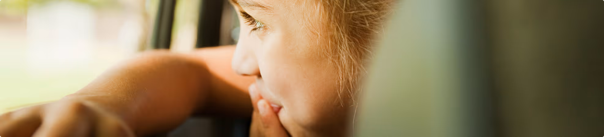 Close-up of a child looking thoughtfully out of a window with their hand near their mouth.