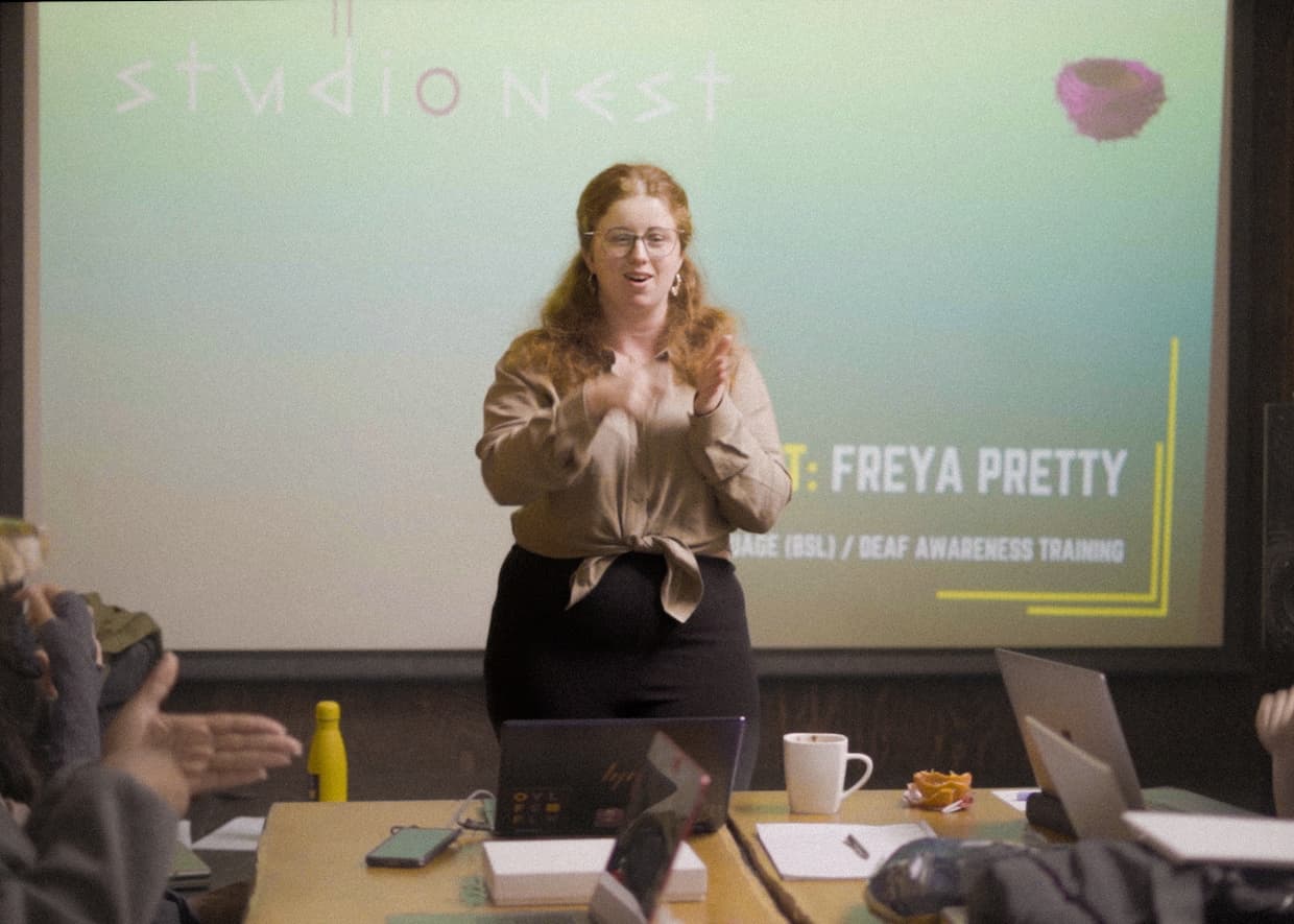 Freya standing in front of a large screen, running a deaf awareness program. She is teaching attendees basic sign language