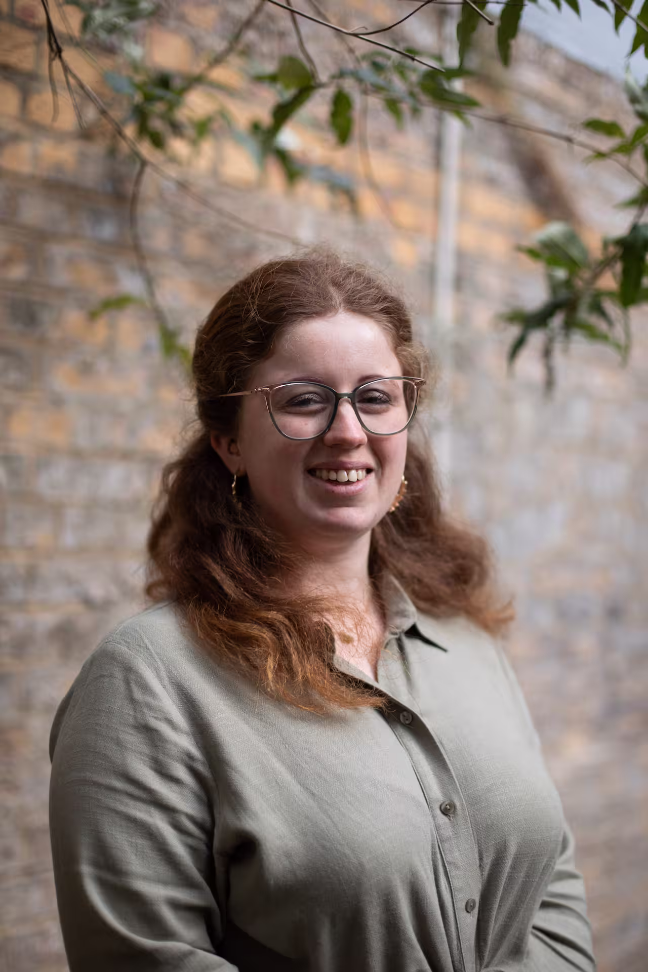 Freya is a white women with wavy red hair, glasses, and light green button-up shirt standing outdoors near a brick wall with green leaves overhead.