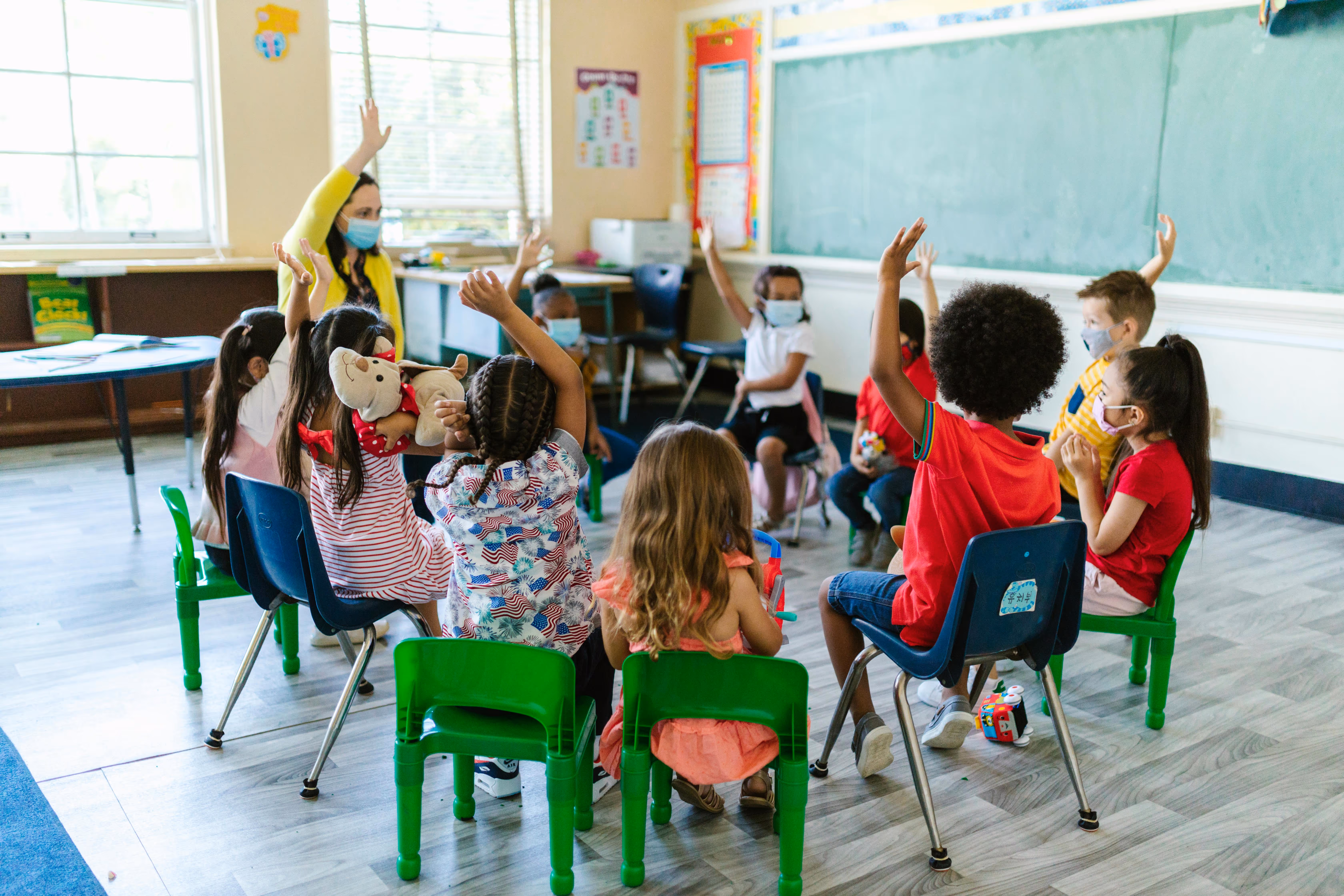 Masked young children sitting in a classroom circle raising their hands with a masked teacher also raising her hand.